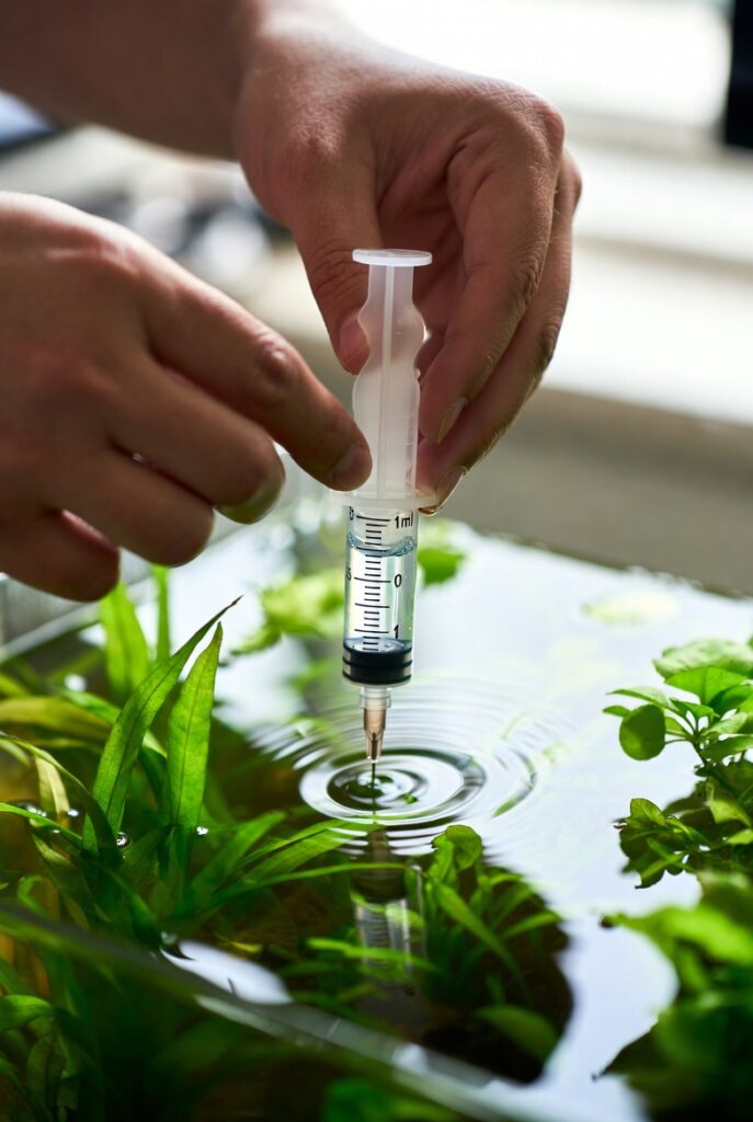 fertilizante líquido aquário plantado. Mãos segurando uma seringa pequena de 1ml dosando fertilizante líquido, prestes a adicionar em um aquário plantado. Plantas verdes visíveis no tanque, iluminação natural limpa, estilo fotorealista